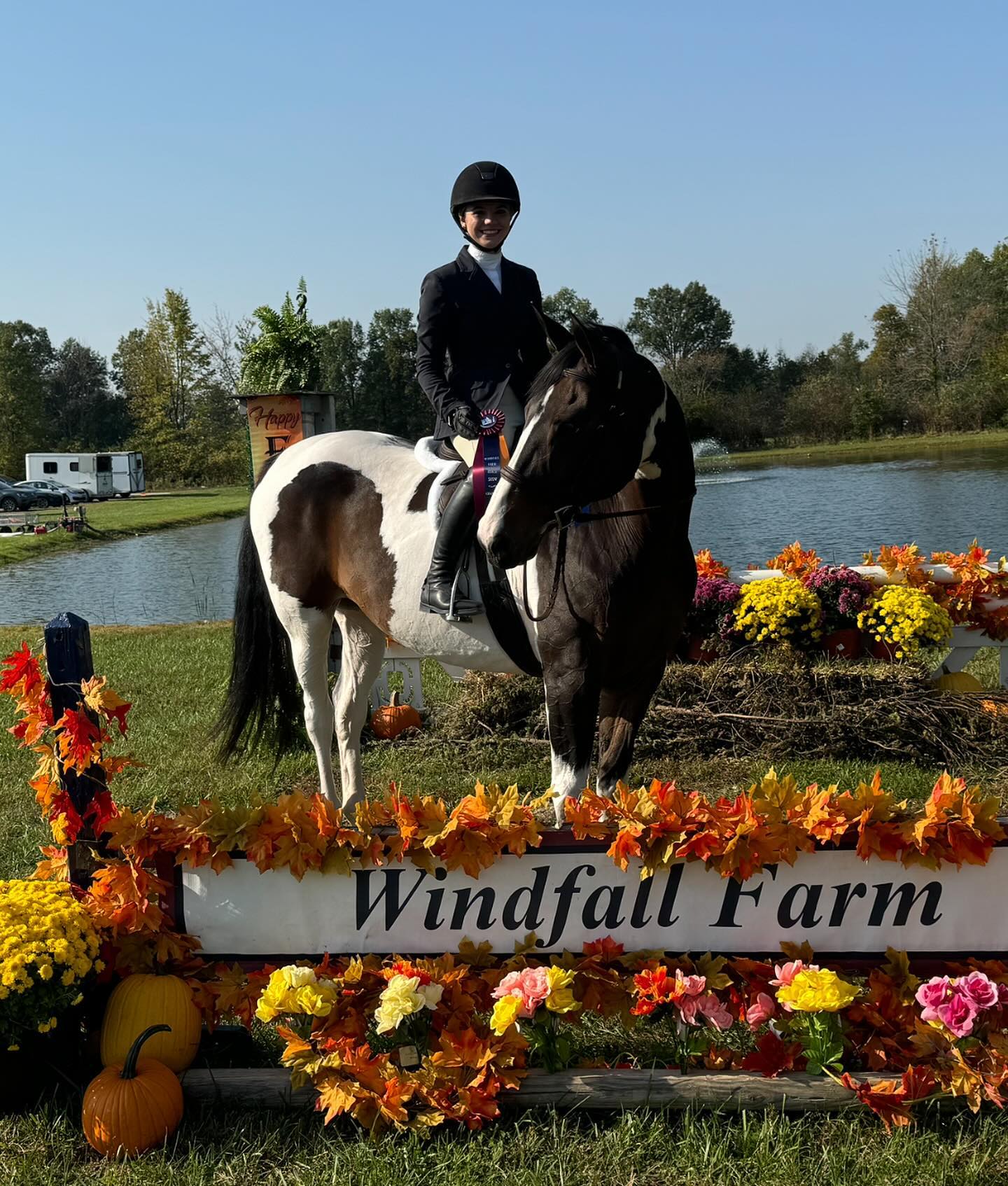 Rylee and SiSi at the Windfall Farm sign with fall decorations, a ribbon, and the lake behind.