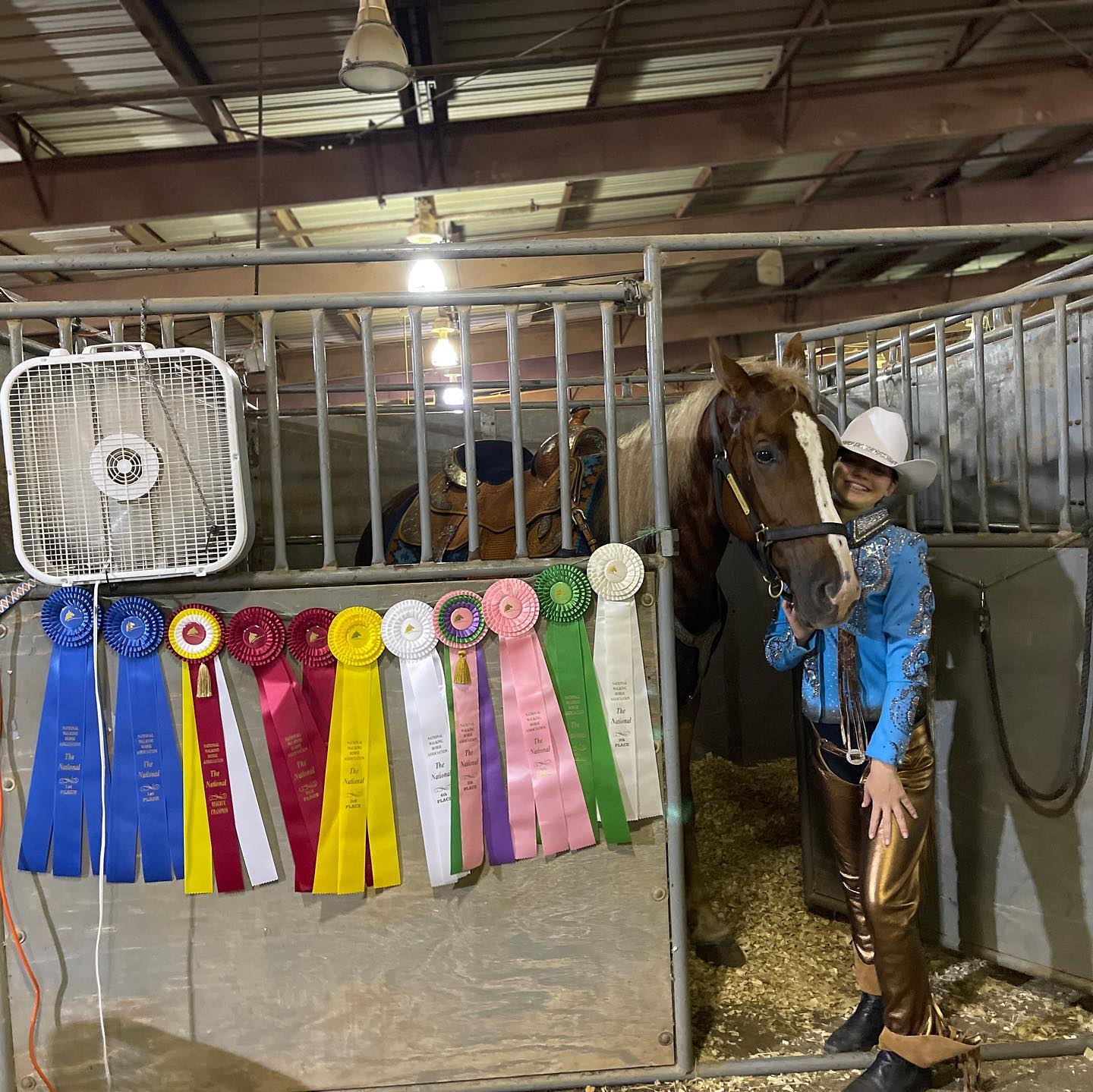 Rylee in a blue western outfit with Sonny beside the NWHA National ribbon wall at the stall.