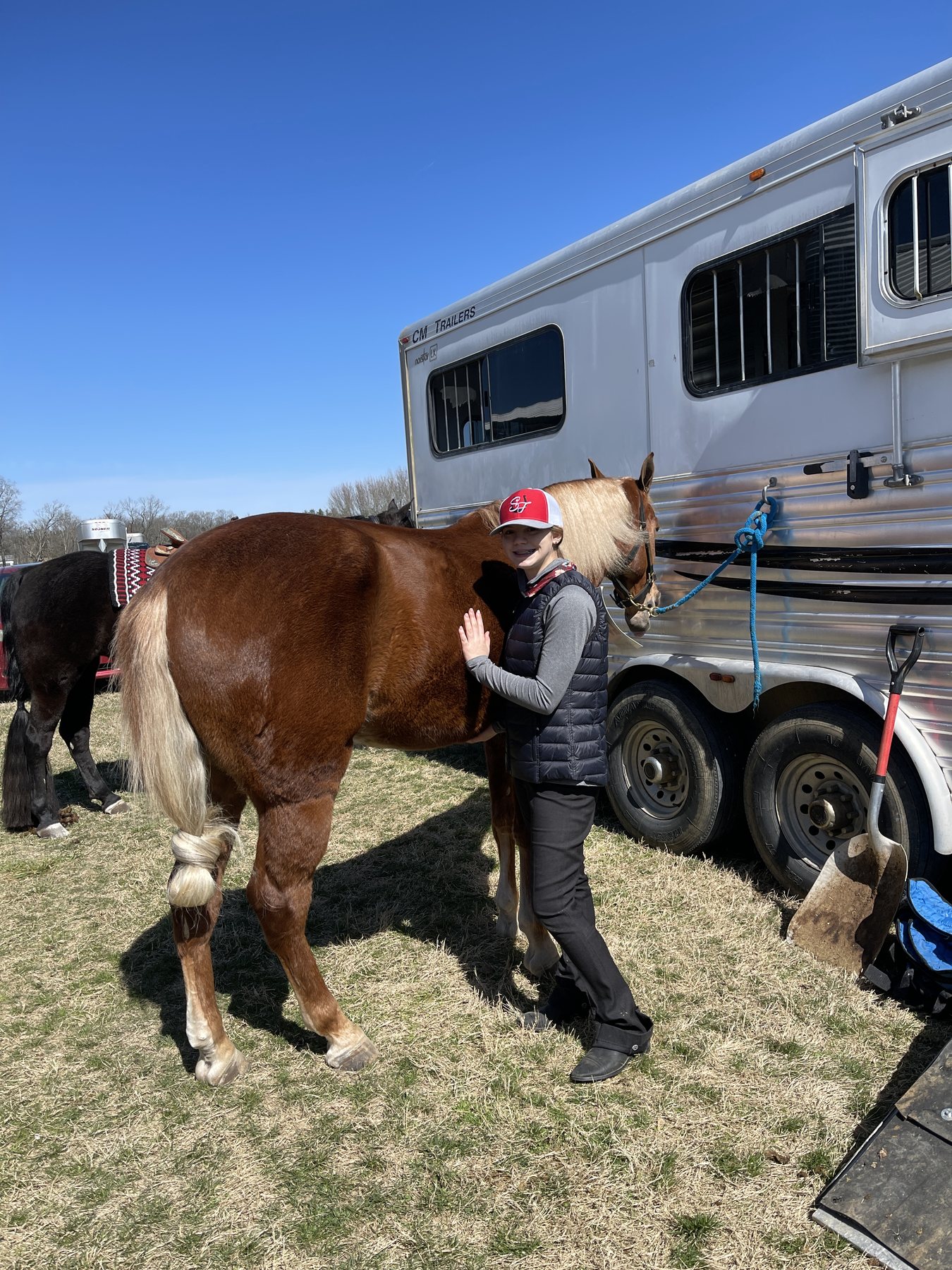 Rylee with her chestnut beside the trailer under a bright blue sky.