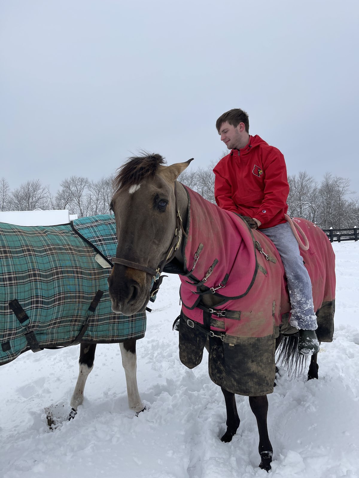Kaleb sitting astride a blanketed horse in deep snow beside a plaid-rugged companion.