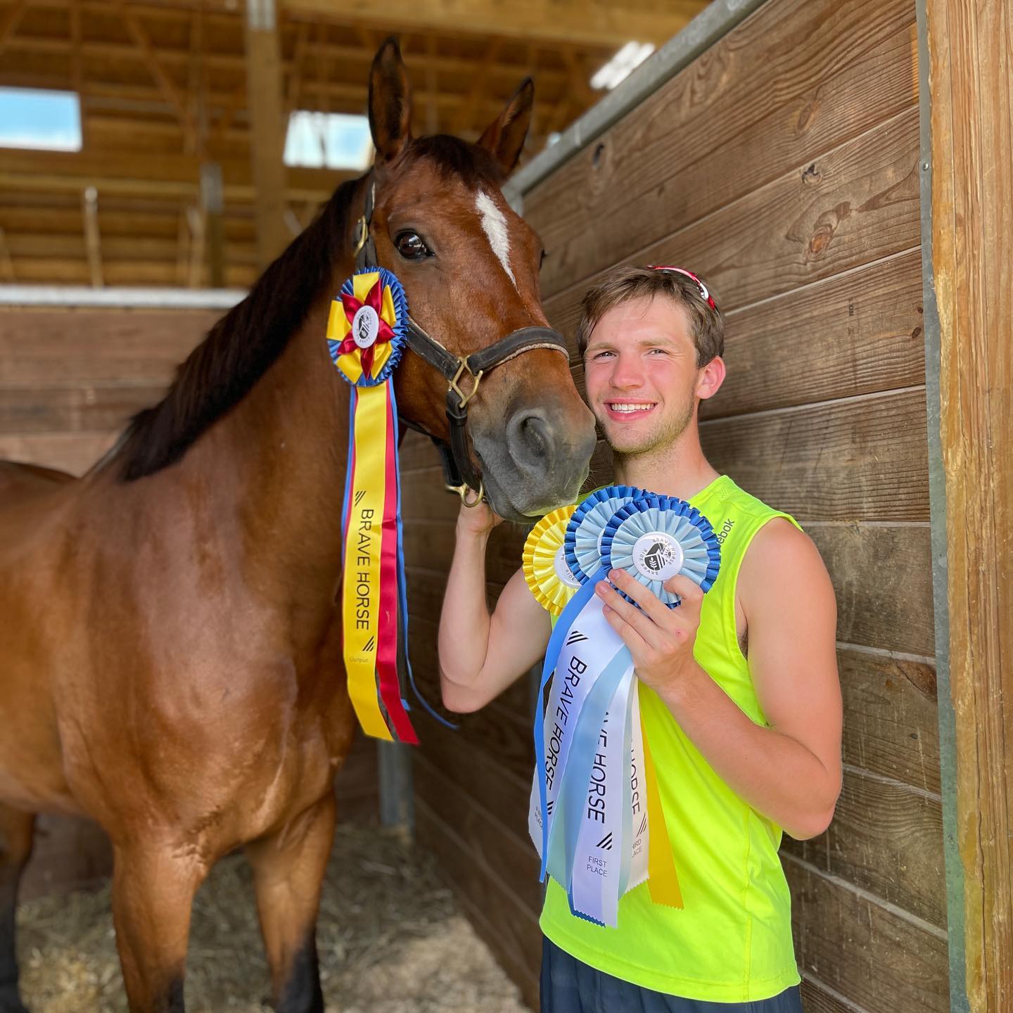 Kaleb with Brave Horse champion ribbons beside a bay in the stall.