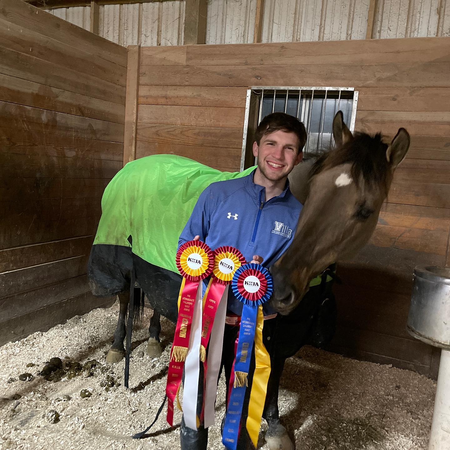 Kaleb's buckskin in a green sheet with KHJA champion rosettes in the stall.