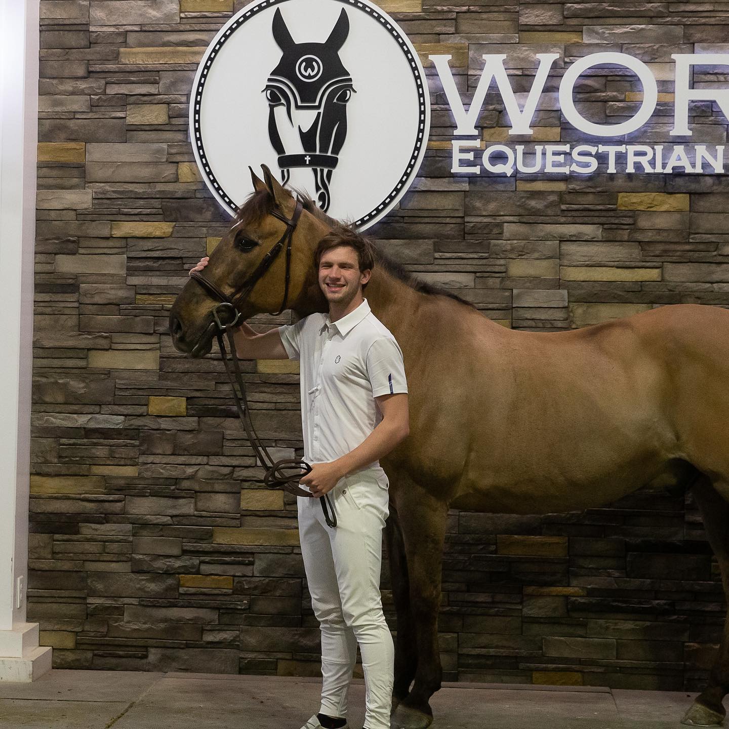 Kaleb in show whites with a buckskin under the World Equestrian Center logo wall.