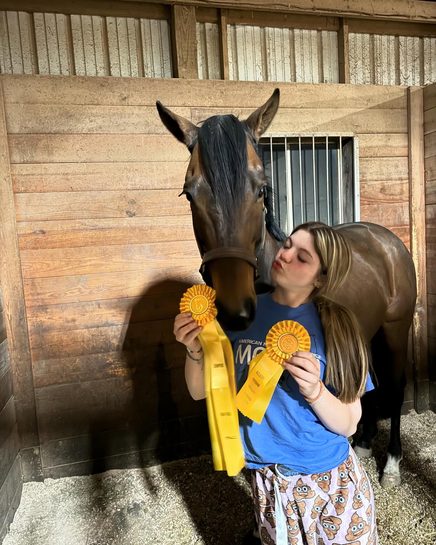 Rylee kissing a bay horse while holding two yellow eventing ribbons in the stall.