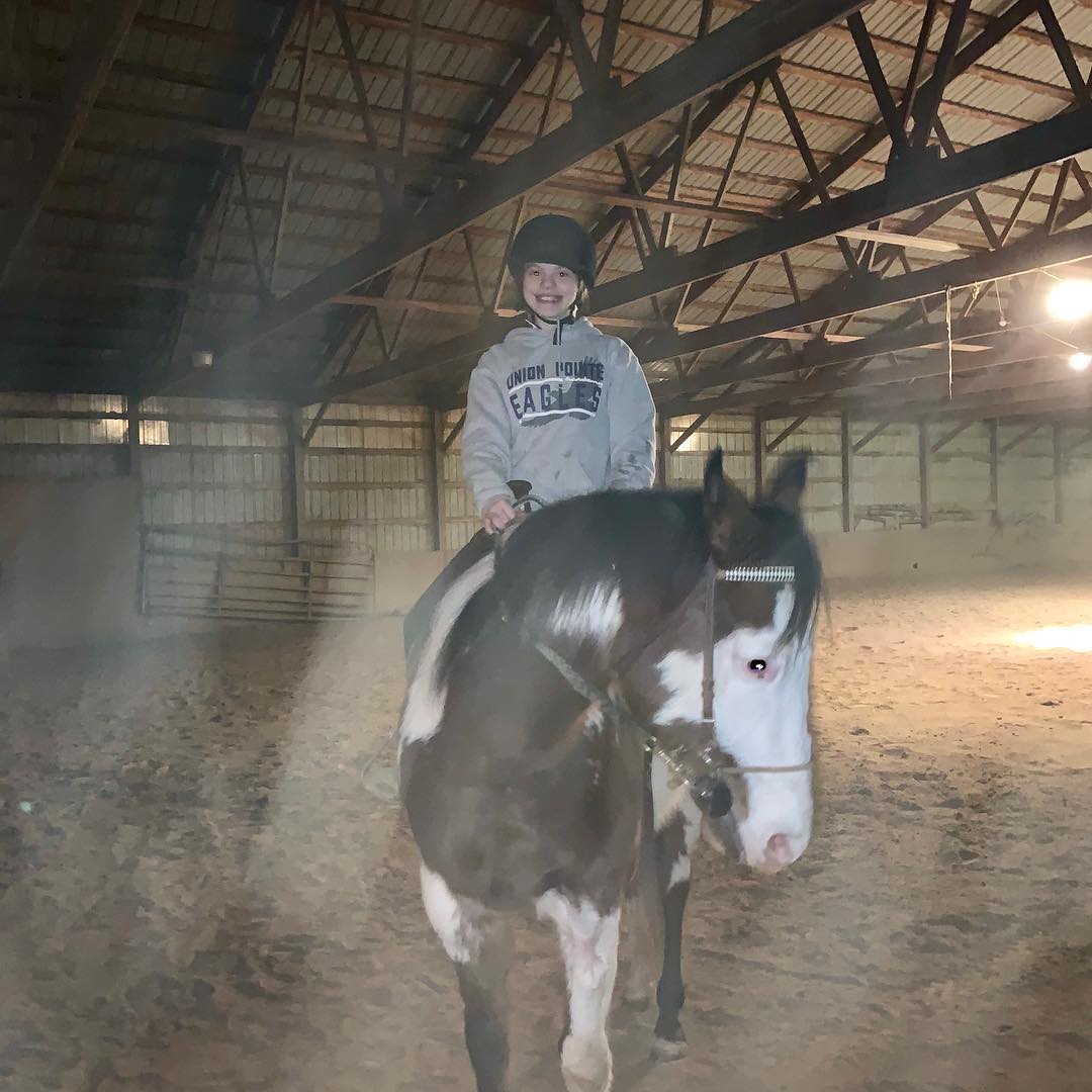 A smiling young Rylee riding a bald-faced, blue-eyed paint bareback in a hazy arena.