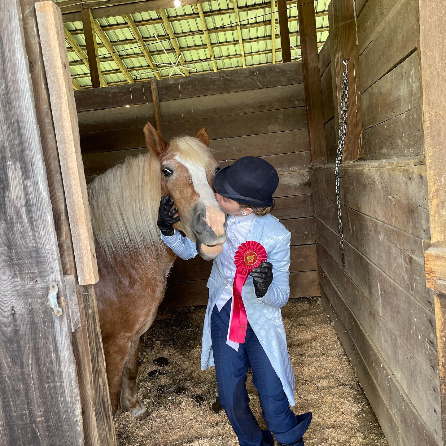 Rylee in a bowler kissing a flaxen Haflinger-type pony with a red ribbon in the stall.