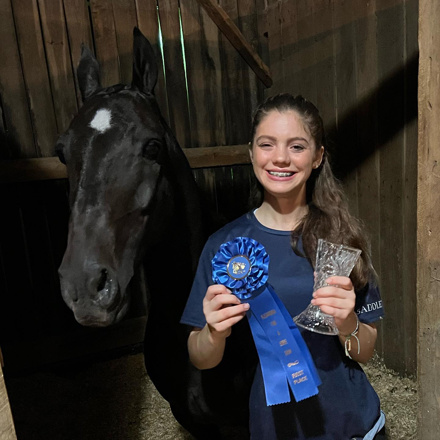 Rylee with a blue ribbon and crystal vase beside a black horse in the stall.