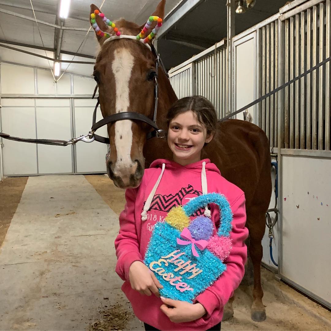 Easter photo of Rylee with a Happy Easter basket and a chestnut wearing pom-pom bunny ears in the barn aisle.
