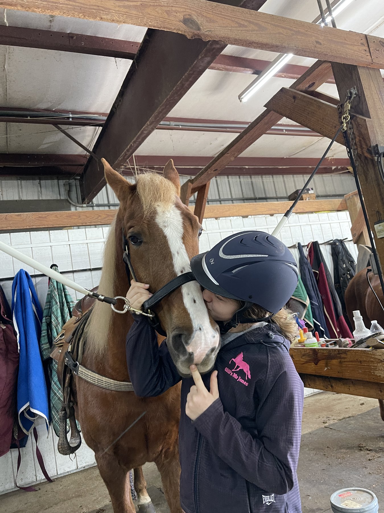Rylee in her riding helmet kissing the muzzle of a flaxen chestnut in the barn aisle.