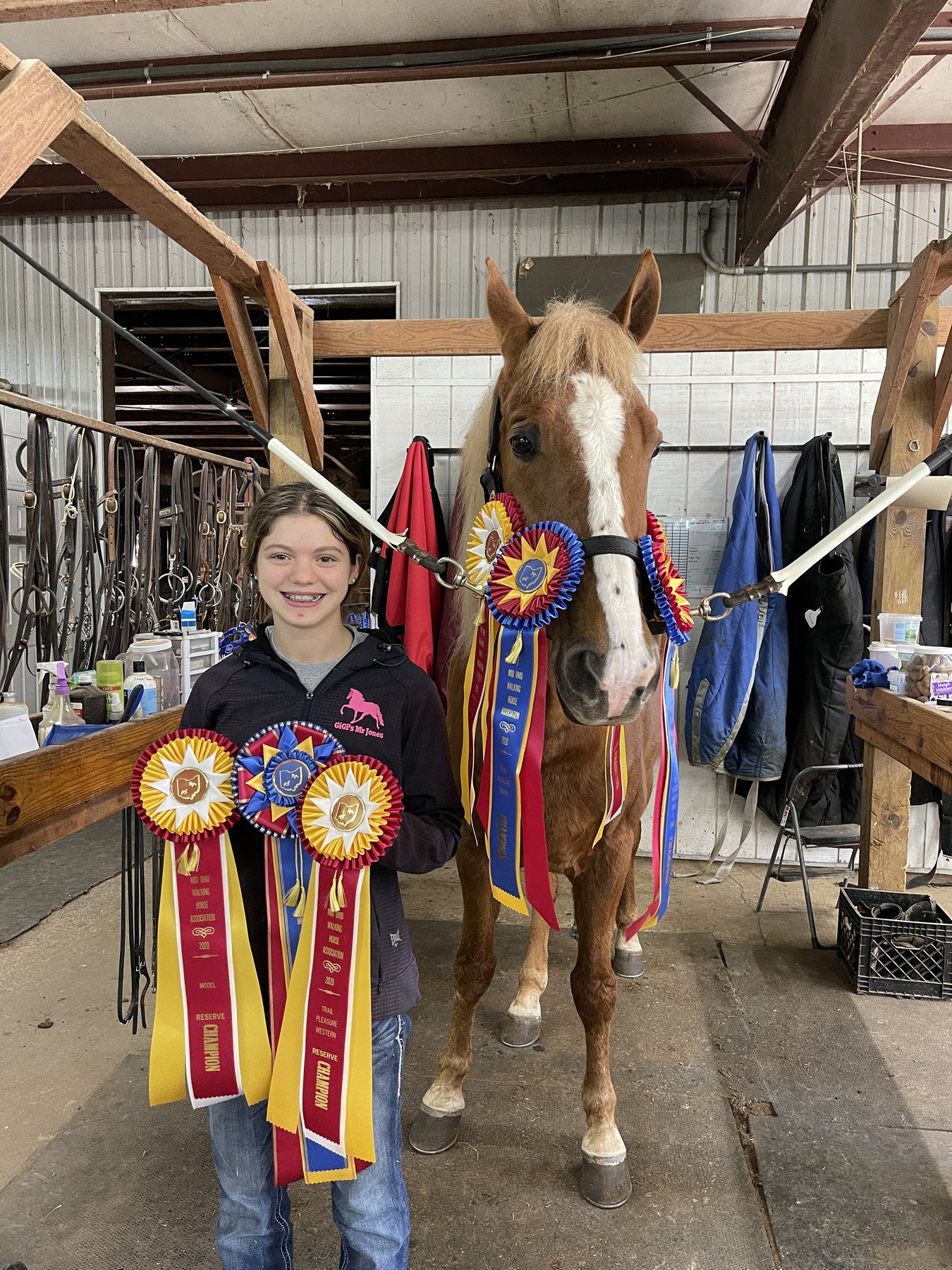 Rylee smiling with an armful of championship ribbons beside a flaxen-maned pony in the barn aisle.
