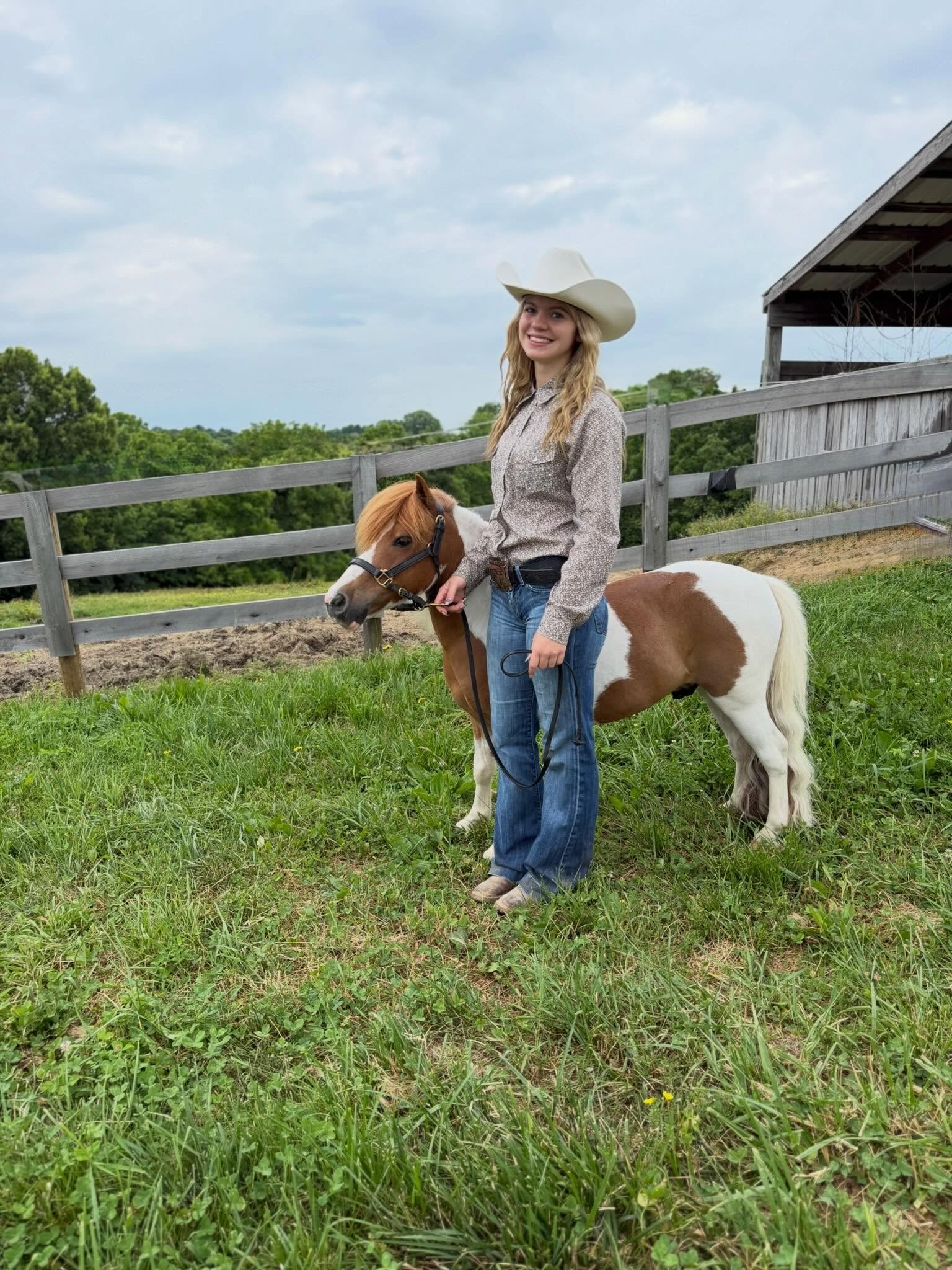 Rylee in a white cowboy hat with a chestnut-and-white miniature horse at the pasture fence.
