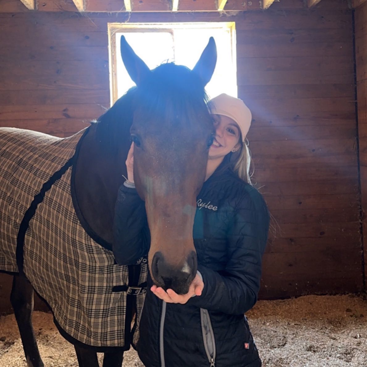 A cozy stall portrait of Rylee in her embroidered jacket with a bay in a plaid blanket.