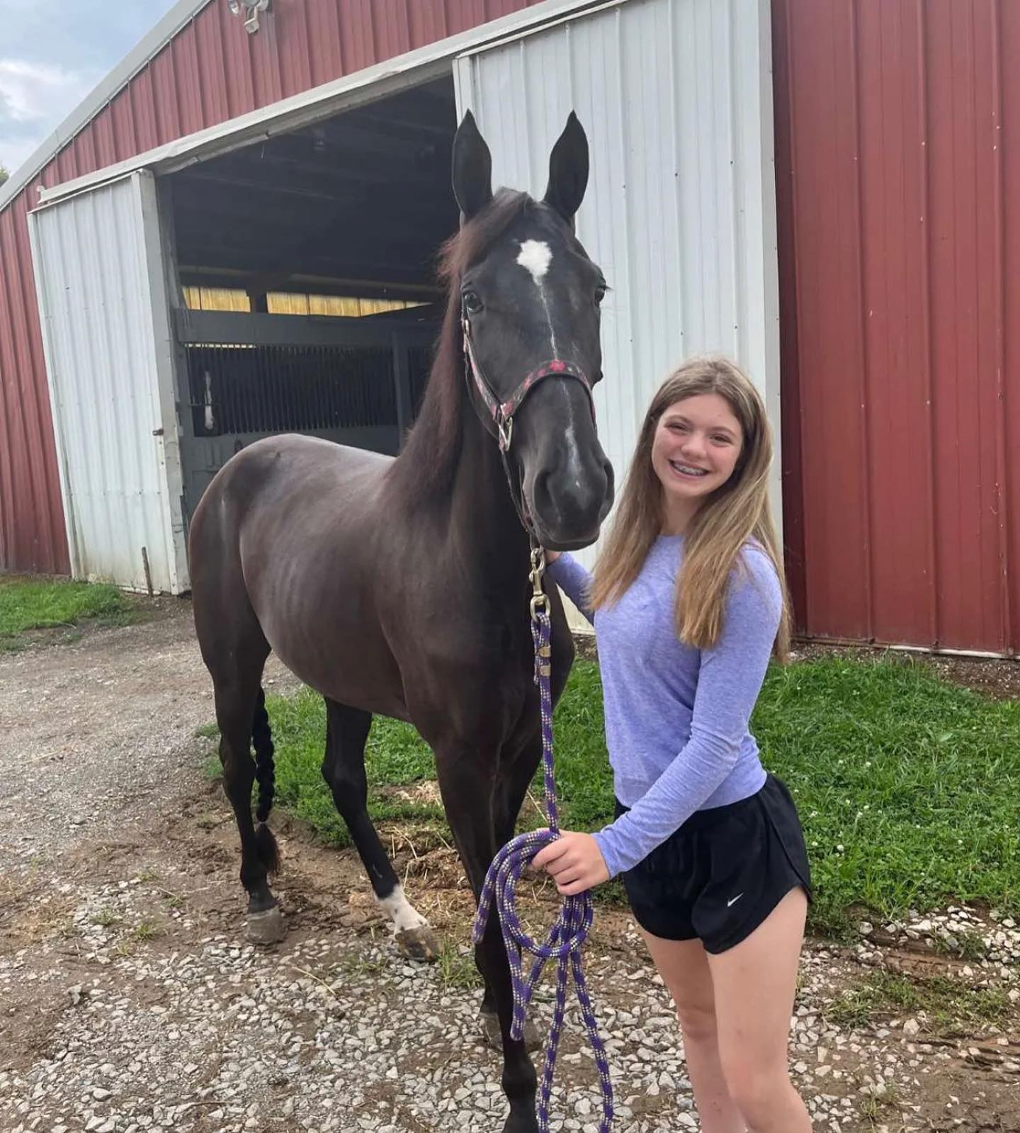 Rylee holding a black horse with a braided tail outside the red barn.