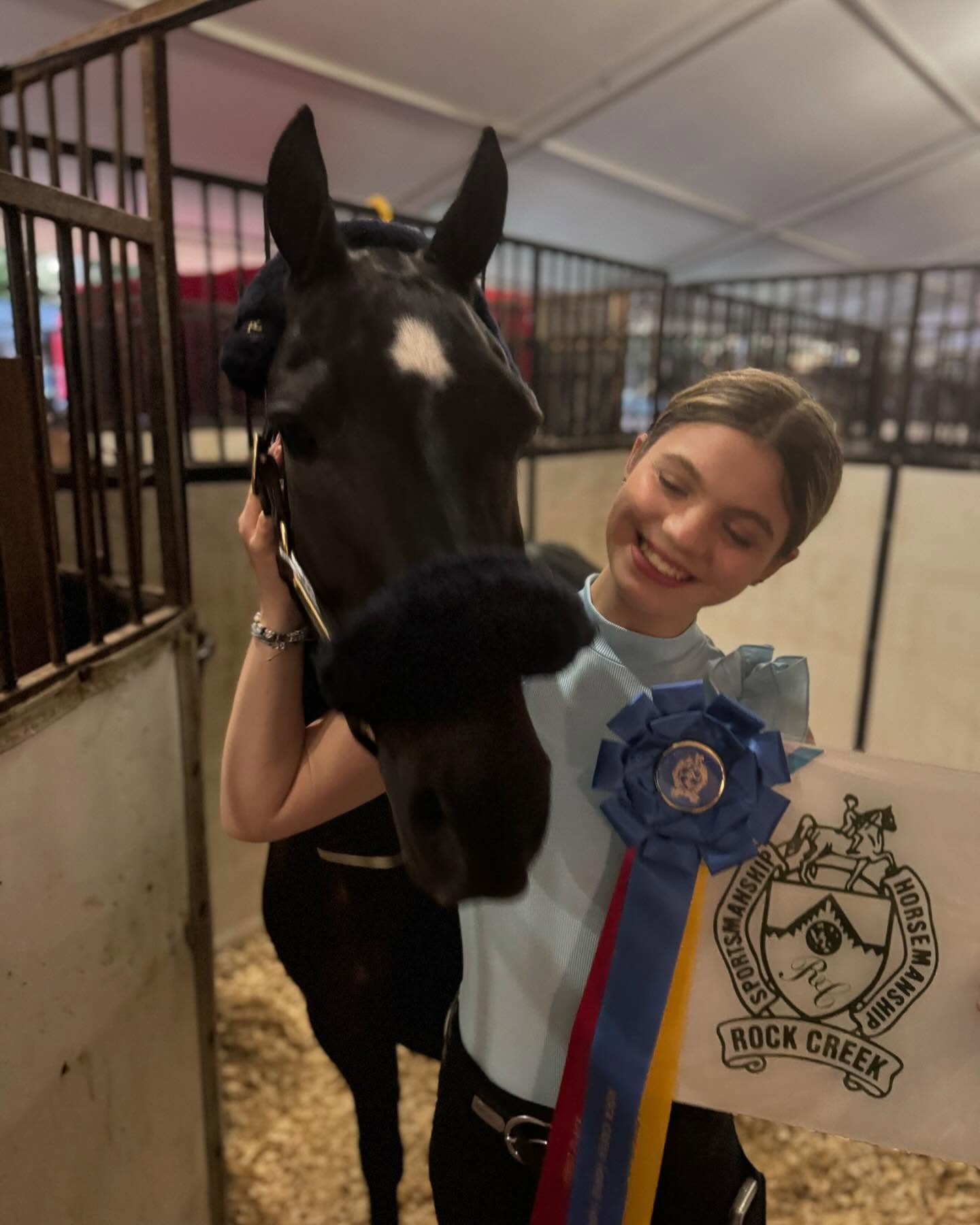Rylee hugging a black horse wearing the Rock Creek sportsmanship blue ribbon in the stabling tent.