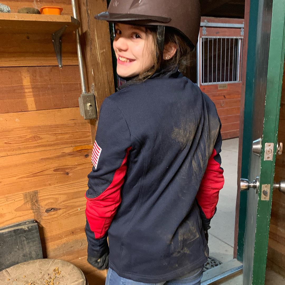A young Rylee in helmet and USA jacket smiling over her shoulder in the barn aisle.