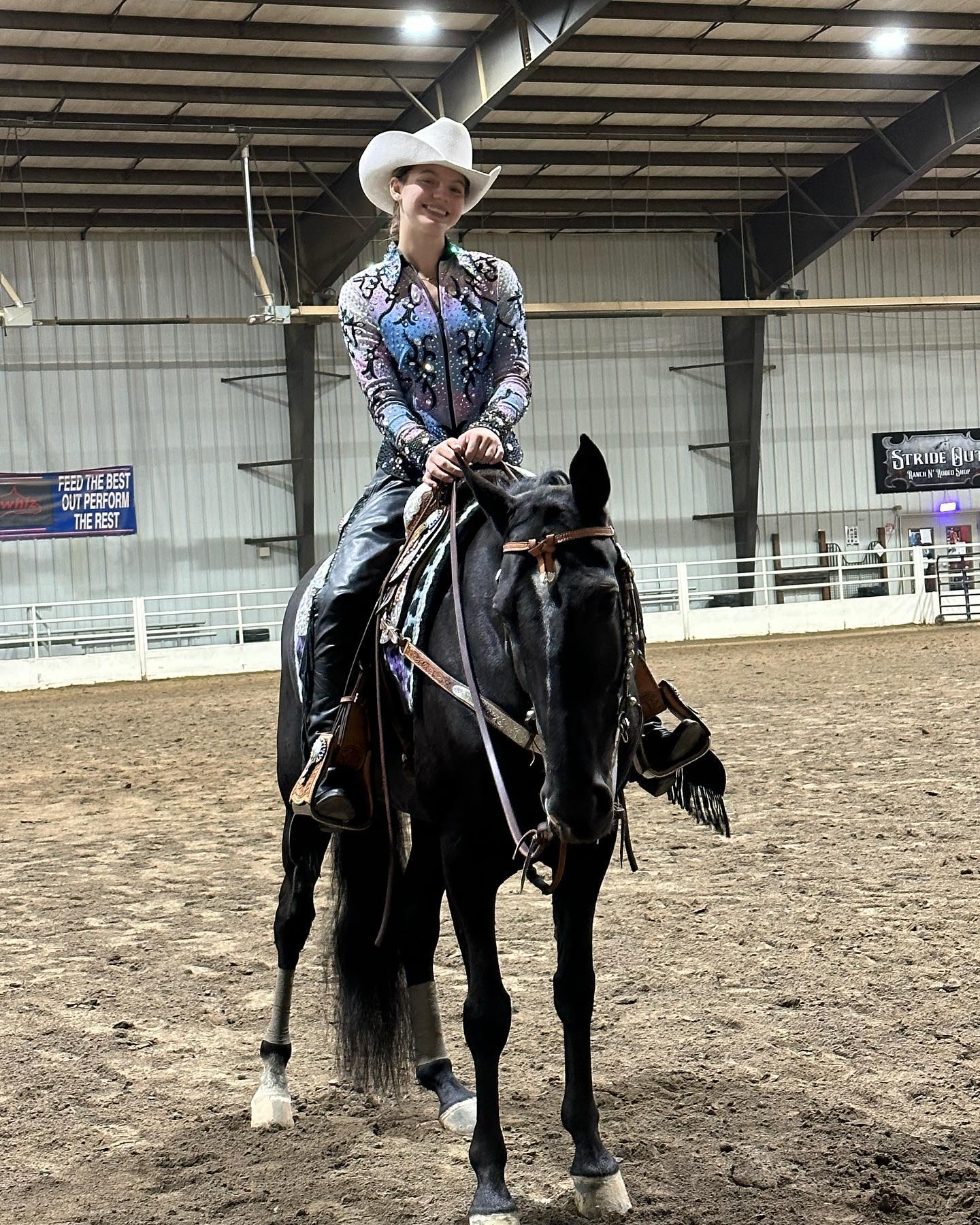 Rylee in a white hat and sparkle shirt showing a black horse western in a sharp indoor arena shot.