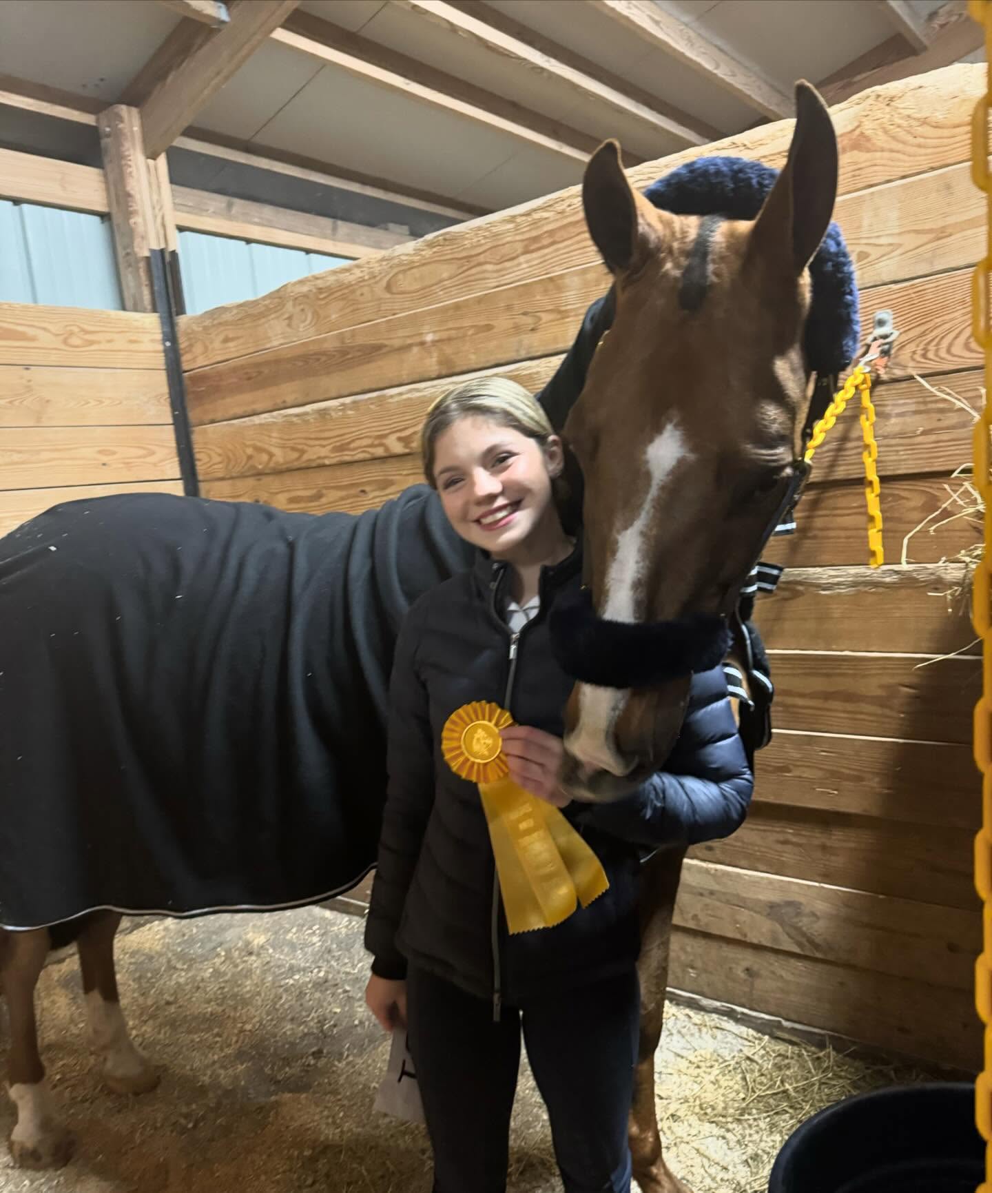 Rylee with a yellow ribbon beside a blanketed, wide-blazed horse in the stall.